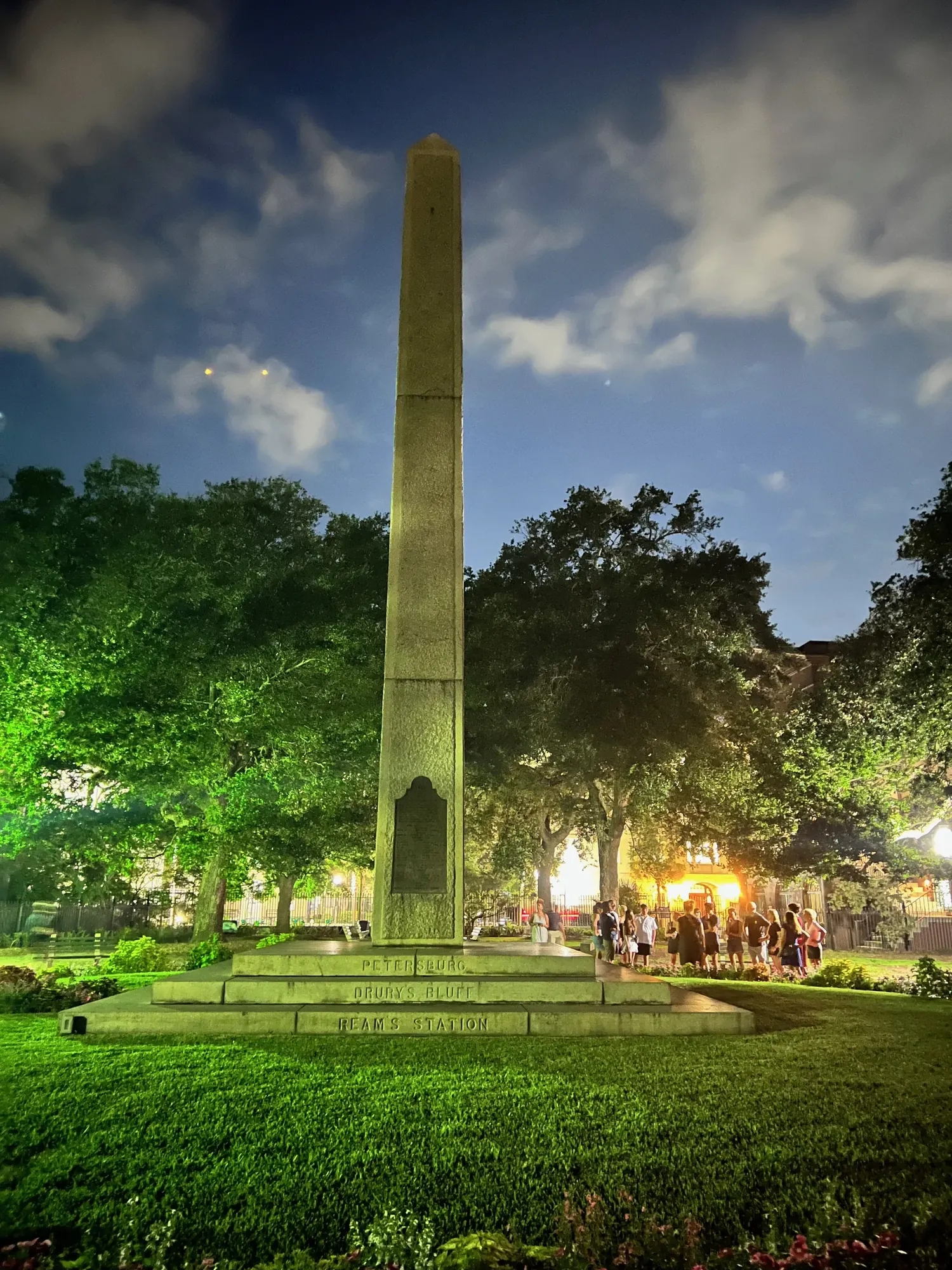 A Confederate memorial obelisk lit at night with a tour group gathered nearby, Charleston