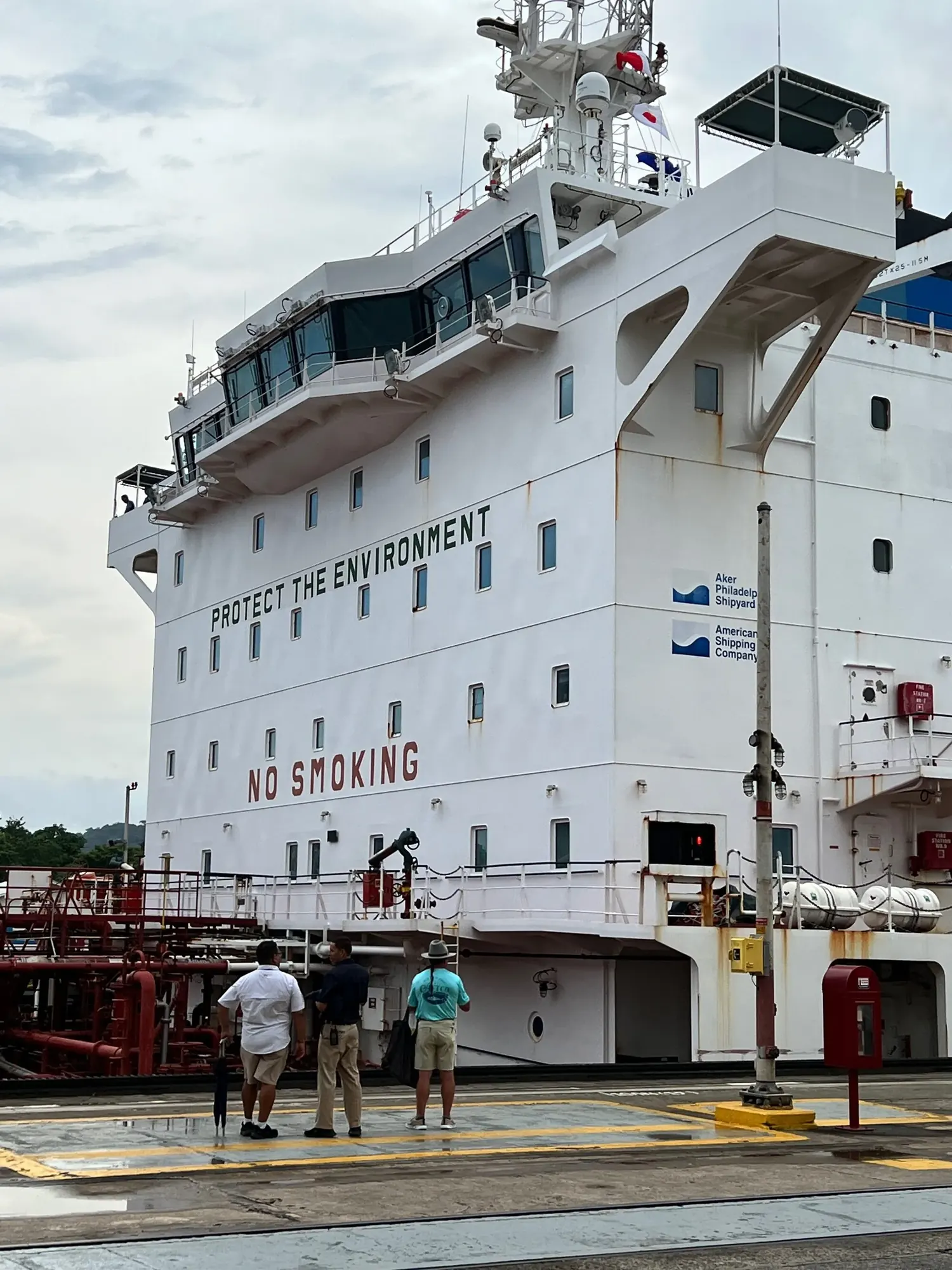 Close view of the white superstructure of a ship inside the Miraflores Locks, marked Protect the Environment and No Smoking, with three visitors watching from the dock below
