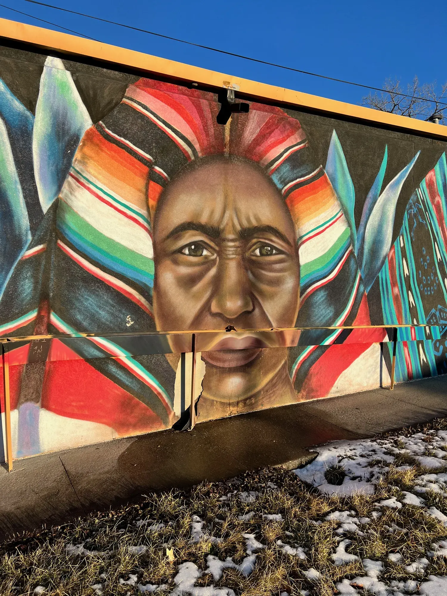 A mural of a Native American figure in a colorful feathered headdress, painted on the side of a building in Des Moines