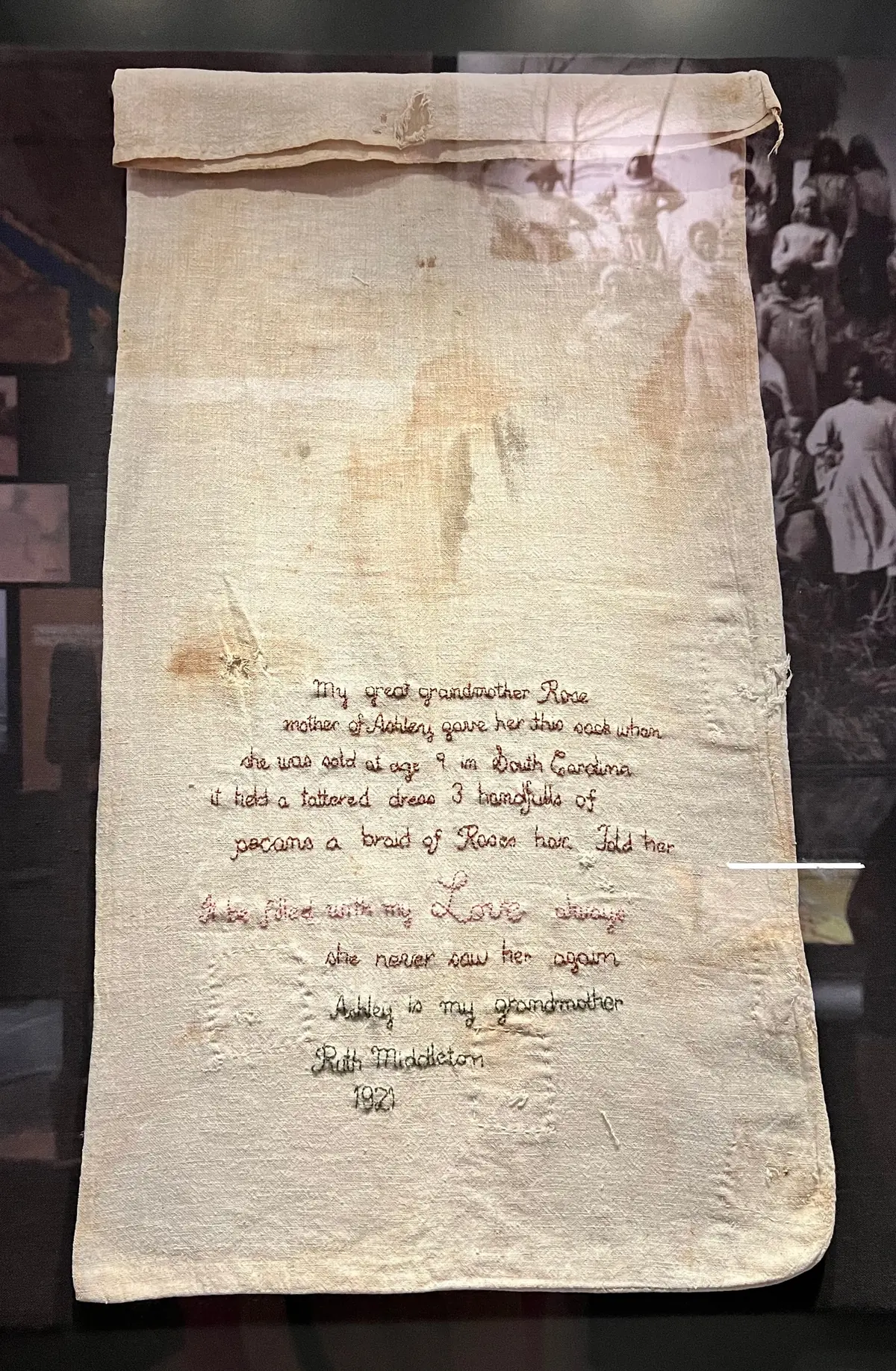 Ashley’s Sack, a worn cotton sack embroidered in 1921 by Ruth Middleton with the story of her great-grandmother Rose and grandmother Ashley