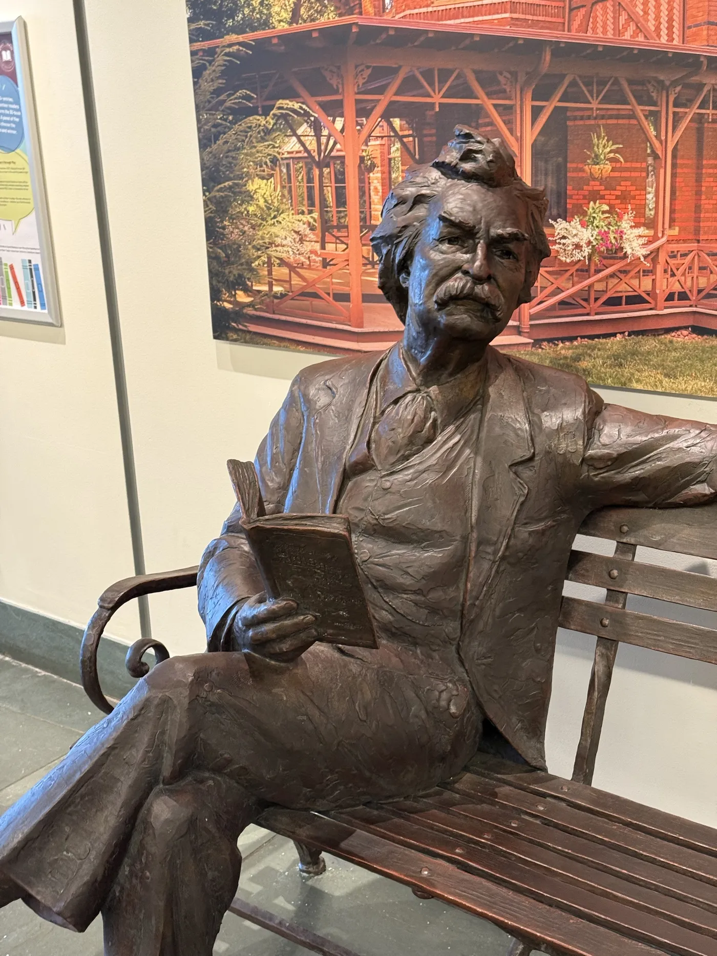 A bronze statue of Mark Twain seated on a bench with a book in his hand, in front of a photograph of the Mark Twain House