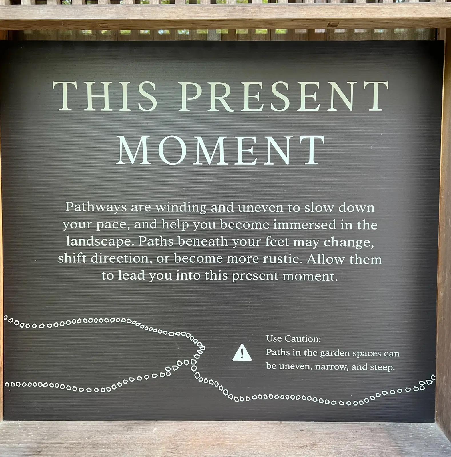 The sign near the Nezu Gate at the Portland Japanese Garden, white lettering on a dark panel reading This Present Moment, with a note that paths in the garden spaces can be uneven, narrow and steep