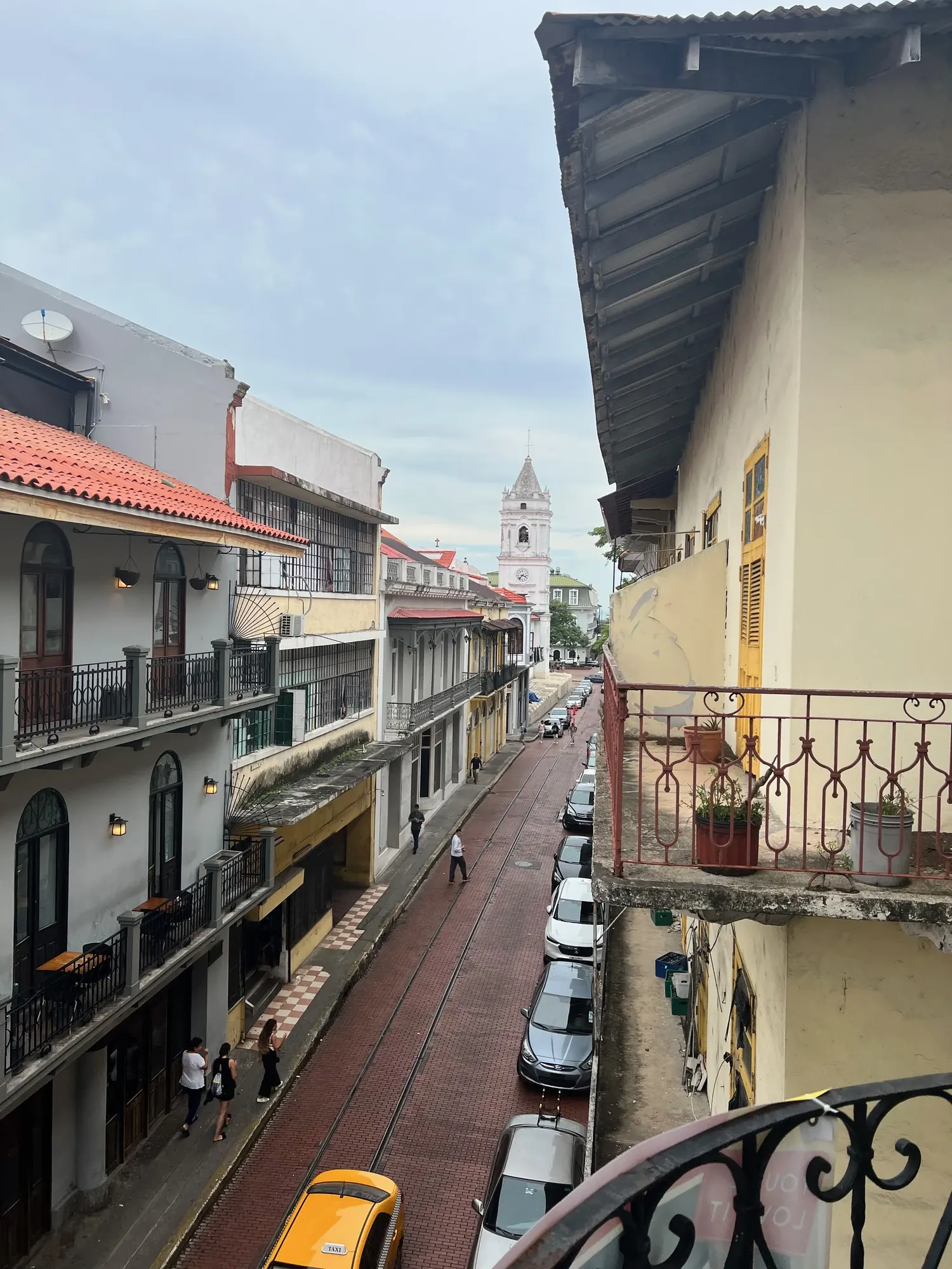 Daytime view down a Casco Viejo street toward a white bell tower, taxi and pedestrians below