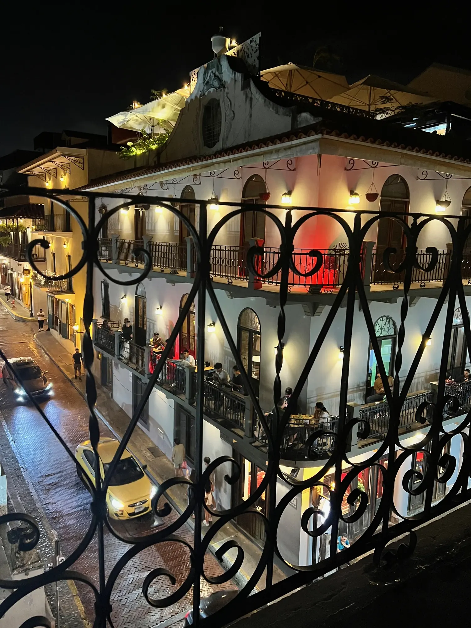 A restaurant across the street lit up at night, seen through a wrought-iron balcony railing